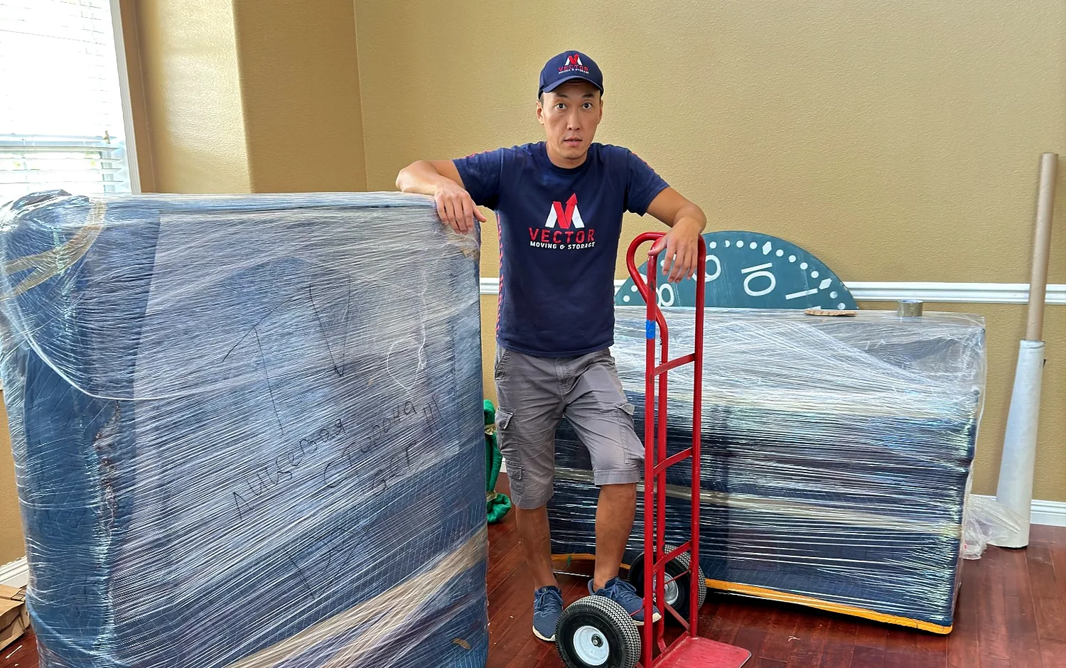 Vector mover standing beside several wrapped furniture pieces and a hand truck